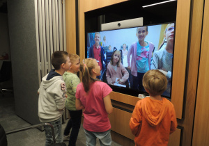 Kilkoro dzieci stoi przed dużym ekranem i rozmawia z dziećmi znajdującymi się w innym pomieszczeniu.
