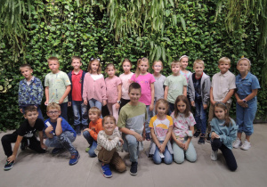 Grupa uczniów stoi na tle ogromnej ściany wyłożonej zielonymi roślinami.