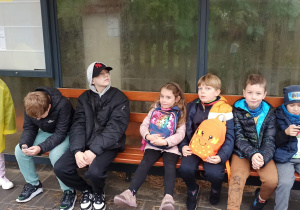 Przystanek autobusowy. Siedmioro dzieci siedzi na ławce pod wiatą przystankową, a dwoje stoi po lewej stronie. Wszyscy wyglądają na zmęczonych ale zadowolonych.