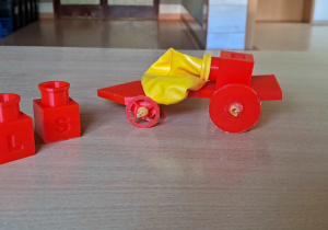Na zdjęciu widać czerwony czterokołowy pojazd z żółtym balonem. Po lewej stronie pojazdu znajdują się dwa dodatkowe sześciany.