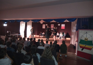 Na scenie stoi trzech uczniów, jeden z nich odgrywa rolę Józefa Piłsudskiego. Na pierwszym planie: widownia oglądająca przedstawienie.