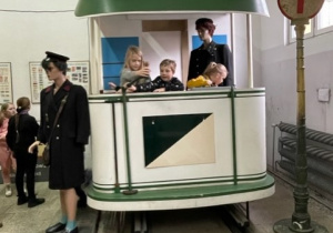 W modelu przedstawiającym dawnego typu wagon tramwajowy znajduje się czwórka uśmiechniętych dzieci. Po prawej stronie zdjęcia przed wagonem stoi staodawny znak p-rzystanku. Po lewej manekin w stroju konduktora.