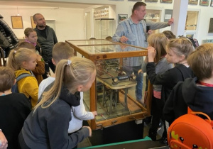 Wokół szklanej gabloty, w której umieszczony jest model napędu tramwajowego stoi grupa dzieci. Przewodnik patrzy w lewo wskazując na coś ręką.