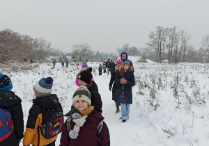 Grupa dzieci i dorosłych na płaskiej zaśnieżonej ścieżce. Na pierwszym planie kilkoro dzieci w różnokolorowych czapkach. Za nimi mężczyzna w niebieskiej kurtce. W oddali reszta grupy.