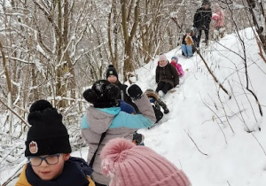 Grupa dzieci w kolorowych ubraniach zbiega i zjeżdża na spodniach z górki po śniegu. W tyle za nimi widać dorosłych na tle ośnieżonych drzew.
