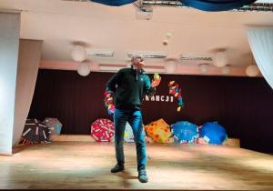 Na zdjęciu żonglujący piłeczkami nauczyciel. W tle na podłodze rozłożone kolorowe parasole.