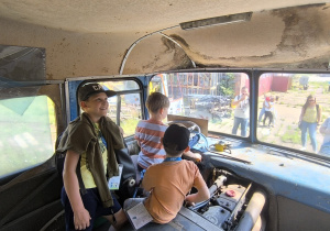 Trzech chłopców, z których dwóch ma na głowie czapki z daszkiem, siedzi w kabinie autobusu muzealnego typu "ogórek", chłopiec bez czapki siedzi tuż przy kierownicy. Za szybą, przed autobusem widać czworo dorosłych.