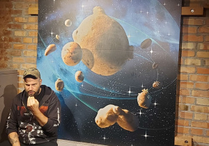 Sala Muzeum Pyry. Na ceglanej ścianie wisi plakat przedstawiający Układ Pyrocentryczny. Po lewej stronie zdjęcia siedzi mężczyzna w czapce z daszkiem. Jest on zwrócony twarzą do dzieci siedzących naprzeciw niego.