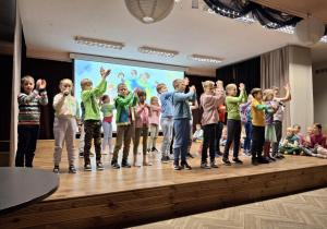 Sala widowiskowa. Na scenie grupa dzieci z uniesionymi nad głowami ramionami klascze w dłonie. dwie dziewczyniki, jedna z prawej, druga z lewej strony sceny trzymają mikrofony. W tle ekran ze zdjęciem.