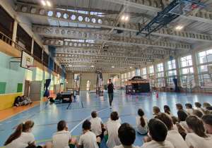 Uczniowie w strojach gimnastycznych, siedzący tyłem w hali sportowej, słuchający zasad objaśnianych przez prowadzącego.