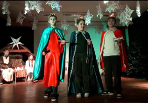 Widok na część sceny, na pierwszym planie stoją uczniowie- trzej królowie trzymający w rękach złoto, kadzidło i mirrę, w tle widać stajenkę wraz z rodziną świętą.