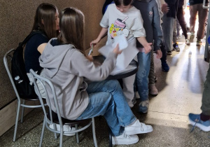 Na zdjęciu widać kolejkę uczniów czekających na odbiór kartek przeznaczonych do głosowania, rozdawanych przy stoliku przez dwie uczennice.