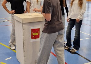 Na zdjęciu widoczny jest uczeń na sali gimnastycznej wrzucający do urny oddany głos. Za urną stoi czwórka uczennic.