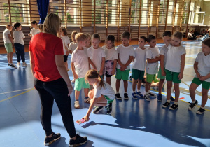 Na zdjęciu widać grupę uczniów w strojach sportowych stojącą przodem do kobiety prowadzącej zajęcia na sali gimnastycznej.