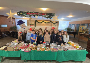 Hol główny szkoły. Na pierwszym planie grupa uczniów, a przed nimi stoły zastawione słodkimi ciastami.