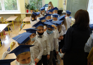Uczniowie z klasy 1a ubrani na galowo, w biretach na głowach, ustawiają się w rzędzie przed Wychowawczynią.