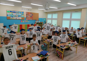 Uczniowie stoją przy ławkach w sali lekcyjnej. Pokazują białe koszulki z nadrukownym imieniem i numerem.