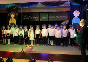 Uczniowie klasy 1c ubrani na galowo, w biretach na głowach, stoją z Panem Dyrektorem na scenie.