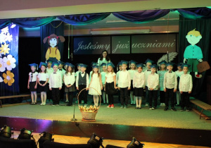 Uczniowie klasy 1c ubrani na galowo, w biretach na głowach, stoją z Wychowawcą na scenie.
